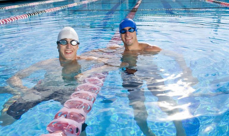 Due nuotatori sorridenti in piscina con cuffie e occhialini.